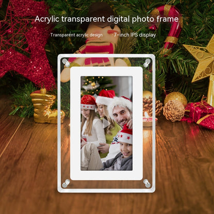 Acrylic transparent digital photo frame placed on a wooden surface with Christmas decorations around it, including red stars, pine cones, and gift boxes. The frame has a transparent acrylic design and features a 7-inch IPS display showcasing a photo of three people wearing Christmas hats.
