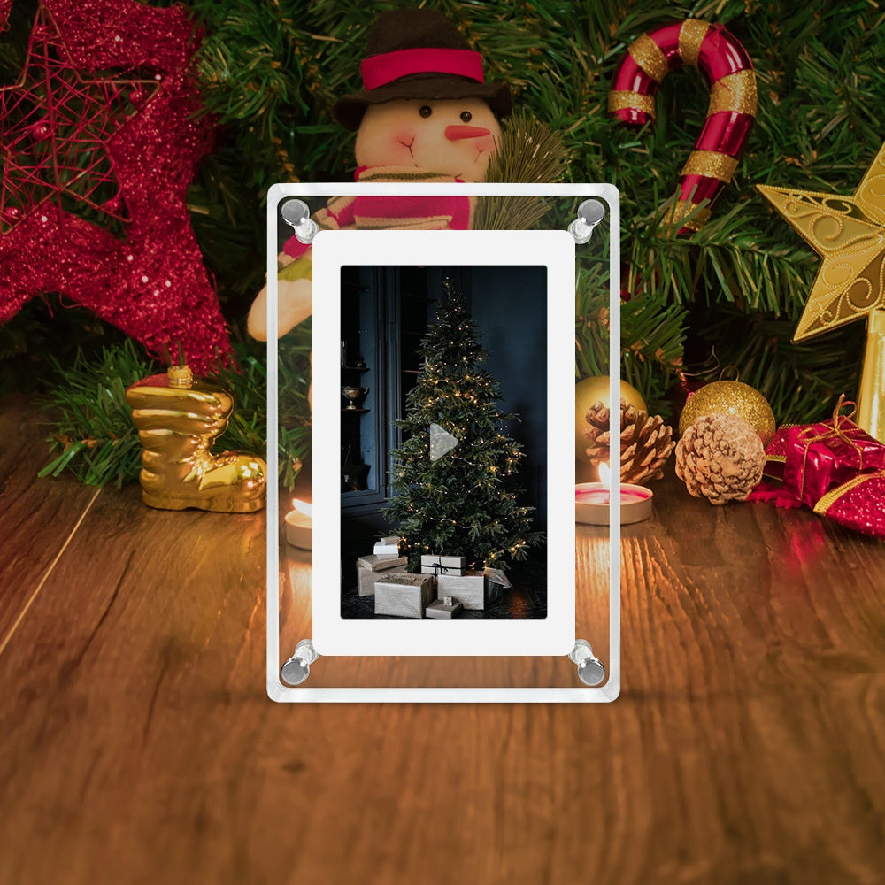 A transparent acrylic digital photo frame displayed on a wooden surface, surrounded by festive Christmas decor, including a snowman figure, candy canes, and pine branches. The frame's screen shows a video play button overlaid on an image of a lit Christmas tree with wrapped presents underneath, suggesting that it is a video player rather than a static photo frame.